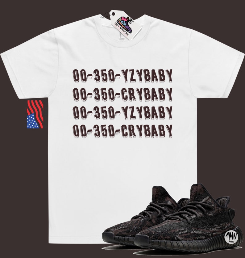 Puede incluir: Camiseta blanca con el texto "00-350-YZYBAY 00-350-CRYBABY 00-350-YZYBAY 00-350-CRYBABY" impreso en marr&oacute;n. Un par de zapatillas negras y marrones con estampado de camuflaje se muestran debajo de la camiseta.