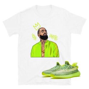 Puede incluir: Una camiseta blanca con una ilustraci&oacute;n de dibujos animados de un hombre que lleva una chaqueta verde ne&oacute;n y una cadena de oro. El texto "HUSTLE" est&aacute; escrito en verde ne&oacute;n sobre la cabeza del hombre. Una corona y un signo de d&oacute;lar est&aacute;n por encima del texto. Un par de zapatillas de deporte verde ne&oacute;n y negras est&aacute;n debajo de la camiseta.
