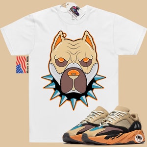 May include: White t-shirt with a cartoon illustration of a dog's face wearing a spiked collar. The dog has brown eyes, a brown nose, and a brown mouth. The t-shirt is paired with a pair of tan, black, and blue sneakers with the text "1MN" on the side.