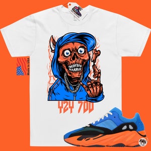 May include: White t-shirt with a cartoon illustration of a skull wearing a blue hoodie and a backwards hat. The skull is making a rock and roll hand gesture. The text "424 700" is printed below the skull. A pair of orange and blue sneakers are shown below the t-shirt.