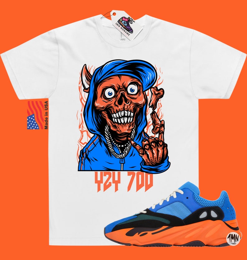 May include: White t-shirt with a cartoon illustration of a skull wearing a blue hoodie and a backwards hat. The skull is making a rock and roll hand gesture. The text "424 700" is printed below the skull. A pair of orange and blue sneakers are shown below the t-shirt.