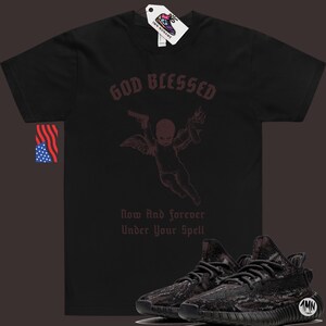 May include: Black t-shirt with a graphic of a cherub holding a gun and a torch. The text "God Blessed" is above the cherub and "Now And Forever Under Your Spell" is below. A pair of black and grey sneakers are in the foreground.