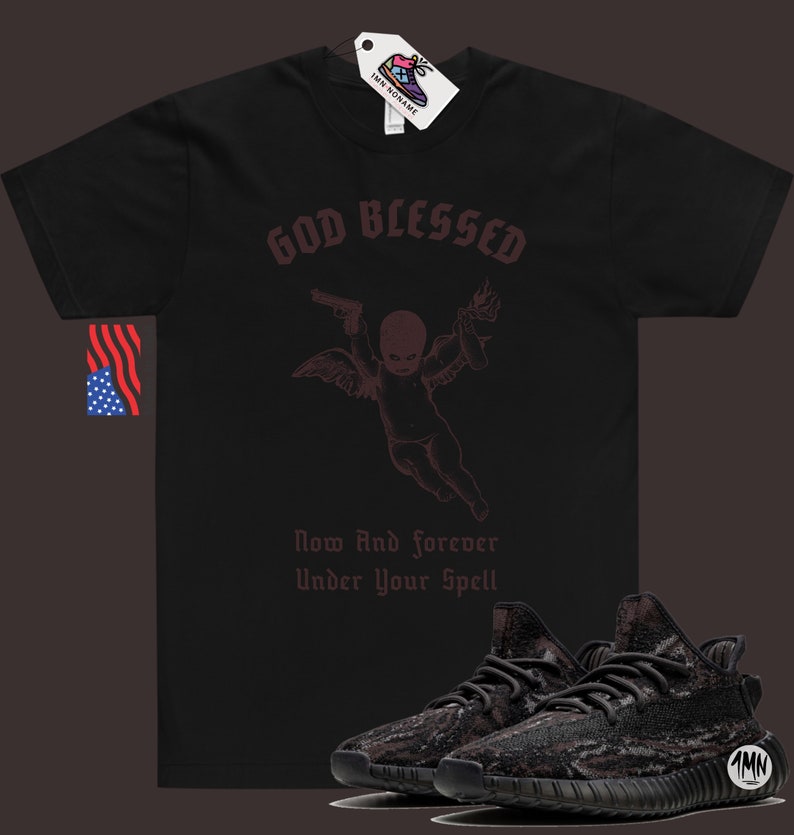 May include: Black t-shirt with a graphic of a cherub holding a gun and a torch. The text "God Blessed" is above the cherub and "Now And Forever Under Your Spell" is below. A pair of black and grey sneakers are in the foreground.