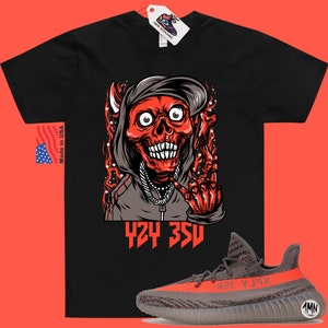 Może przedstawiać: Czarny T-shirt z czerwoną i szarą ilustracją czaszki w kapturze i łańcuchu. Tekst "424 350" jest nadrukowany pod ilustracją. Para szarych i pomarańczowych but&oacute;w Yeezy Boost 350 V2 jest pokazana pod T-shirtem.