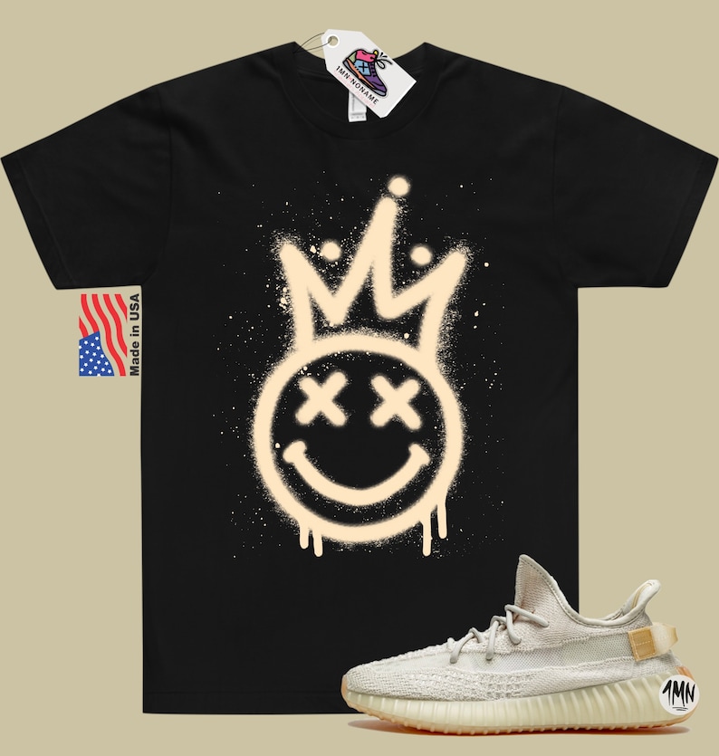 Puede incluir: Camiseta negra con un dise&ntilde;o de pintura en aerosol blanco de una corona y una cara sonriente con X para los ojos. Un par de zapatillas de deporte de color beige claro con suela marr&oacute;n y una peque&ntilde;a etiqueta que dice "1MN" est&aacute;n en la esquina inferior derecha de la imagen.