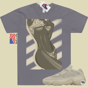 Puede incluir: Una camiseta gris con un gr&aacute;fico de una mujer en traje de ba&ntilde;o. La camiseta tiene una etiqueta que dice "1MN" y las palabras "Made in USA". Tambi&eacute;n se muestra un par de zapatillas beige con una parte superior de malla y una suela gruesa.