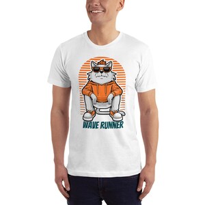 May include: White t-shirt with a cartoon cat wearing sunglasses and an orange hoodie, sitting on a white chair. The cat has a red headband and the text "WAVE RUNNER" is printed below the cat.