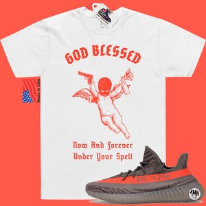Puede incluir: Camiseta blanca con texto rojo que dice "God Blessed" y una ilustraci&oacute;n roja de un querub&iacute;n con alas que sostiene una pistola y un c&oacute;ctel Molotov. El texto "Now And Forever Under Your Spell" est&aacute; debajo de la ilustraci&oacute;n. Un par de zapatillas grises y naranjas est&aacute;n debajo de la camiseta.