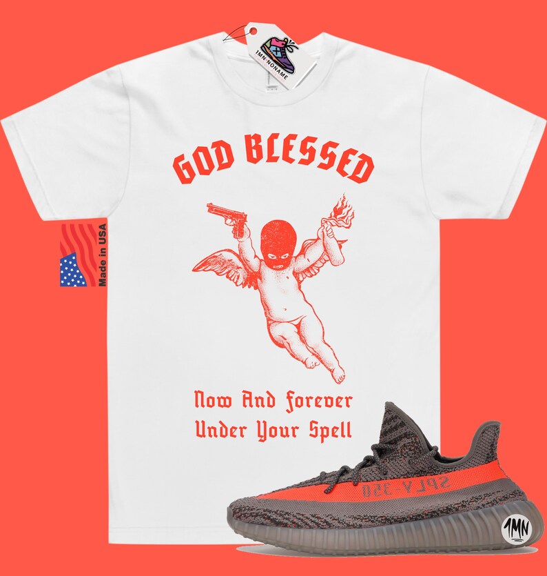 Puede incluir: Camiseta blanca con texto rojo que dice "God Blessed" y una ilustraci&oacute;n roja de un querub&iacute;n con alas que sostiene una pistola y un c&oacute;ctel Molotov. El texto "Now And Forever Under Your Spell" est&aacute; debajo de la ilustraci&oacute;n. Un par de zapatillas grises y naranjas est&aacute;n debajo de la camiseta.