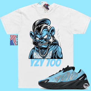 May include: A white t-shirt with a blue cartoon monkey wearing a hat and a chain. The text "YZY 700" is printed below the monkey. A pair of blue and black Yeezy 700 sneakers are shown below the t-shirt.