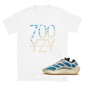 Puede incluir: Camiseta blanca con "700 YZY" impreso en rayas azules y verdes. Un par de zapatillas blancas y azules con suela amarilla.