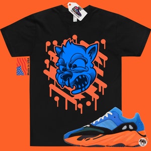 May include: A black t-shirt with a blue cartoon wolf graphic and orange dripping paint. The shirt is paired with blue and orange sneakers with the text "1MN" on the side.