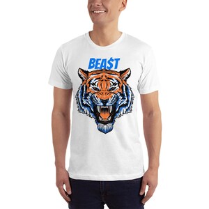 May include: White t-shirt featuring a detailed illustration of a tiger's face with orange and blue accents. The word "BEA$T" is printed above the tiger in blue. The shirt is a short-sleeved crew neck.