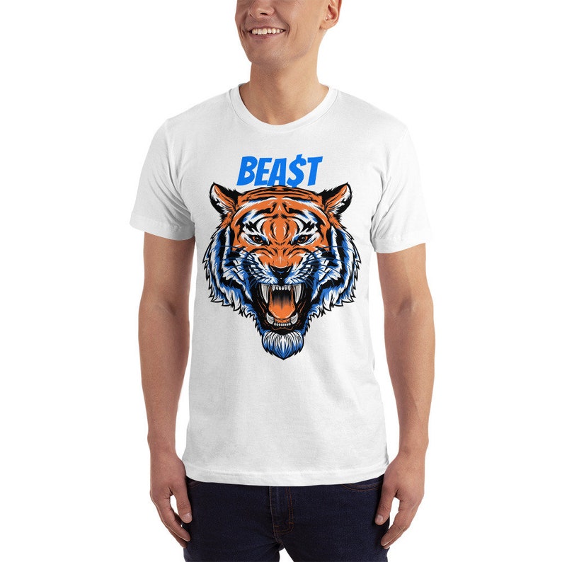 May include: White t-shirt featuring a detailed illustration of a tiger's face with orange and blue accents. The word "BEA$T" is printed above the tiger in blue. The shirt is a short-sleeved crew neck.