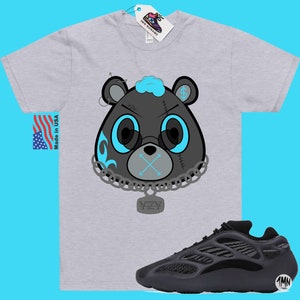 May include: A grey t-shirt featuring a cartoon black bear wearing a chain with a YZY pendant. The bear has blue eyes, a blue mohawk, and a blue mouth with an X over it. The bear is wearing a chain with a YZY pendant. The t-shirt is paired with black Yeezy 700 trainers.