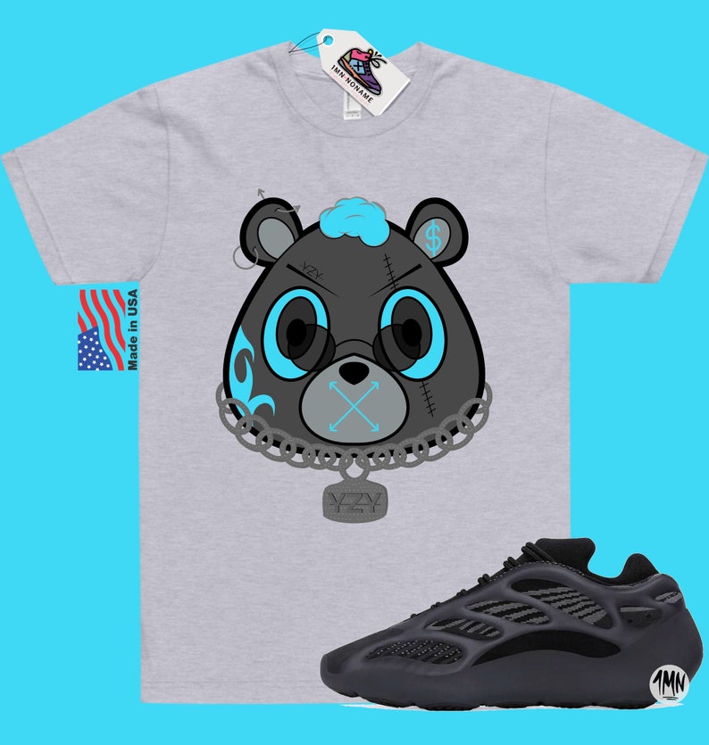 May include: A grey t-shirt featuring a cartoon black bear wearing a chain with a YZY pendant. The bear has blue eyes, a blue mohawk, and a blue mouth with an X over it. The bear is wearing a chain with a YZY pendant. The t-shirt is paired with black Yeezy 700 trainers.