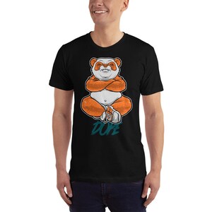 Pu&ograve; includere: Maglietta nera con una grafica di un panda arancione e bianco seduto con le braccia incrociate. Il panda indossa scarpe da ginnastica e il testo "DOPE" &egrave; stampato sotto.