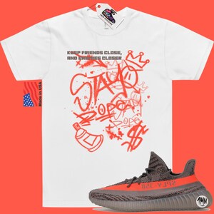 Puede incluir: Camiseta blanca con texto rojo de estilo graffiti que dice "Keep friends close, and enemies closer" y "Stay" con una corona sobre el texto. Un par de zapatillas grises y naranjas se encuentran debajo de la camiseta.