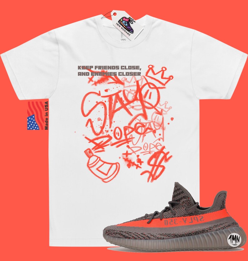 Puede incluir: Camiseta blanca con texto rojo de estilo graffiti que dice "Keep friends close, and enemies closer" y "Stay" con una corona sobre el texto. Un par de zapatillas grises y naranjas se encuentran debajo de la camiseta.