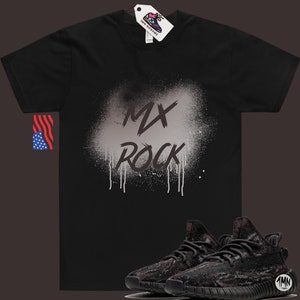以下が含まれることがあります： 白いスプレーペイントのデザインで「MX Rock」と書かれた黒いTシャツ。Tシャツの下には、黒と茶色の迷彩柄のスニーカーが1足あります。