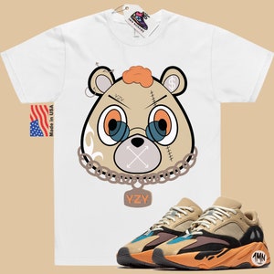 May include: White t-shirt featuring a cartoon bear wearing a chain and a tag that reads "YZY". The bear has a stitched-up face with an X over its mouth. The t-shirt is paired with brown and orange trainers with a black sole and a tag that reads "1MN".