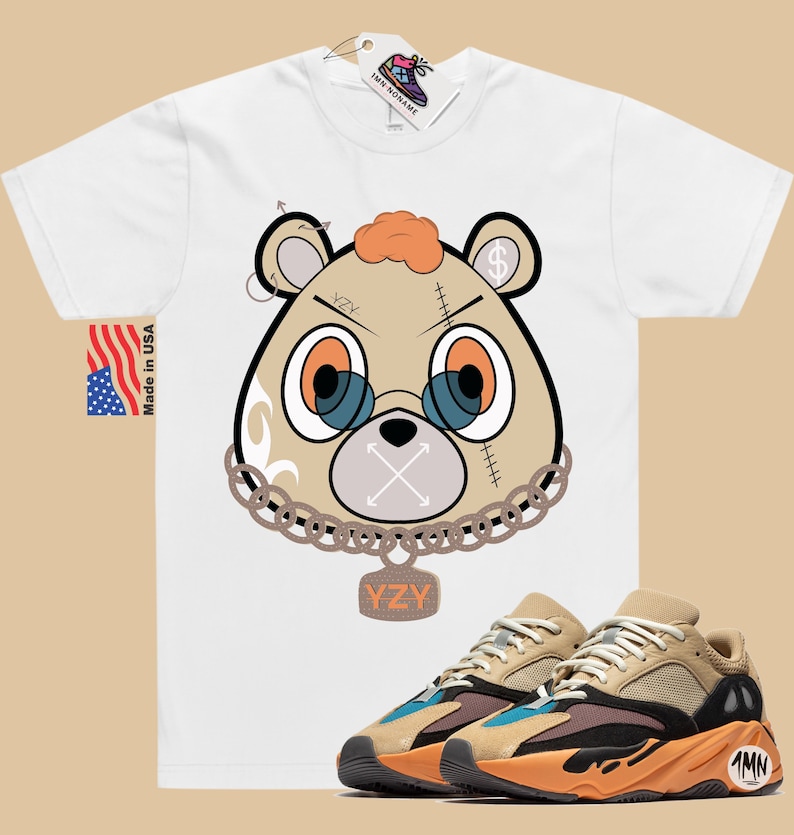 May include: White t-shirt featuring a cartoon bear wearing a chain and a tag that reads "YZY". The bear has a stitched-up face with an X over its mouth. The t-shirt is paired with brown and orange trainers with a black sole and a tag that reads "1MN".