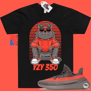Pode incluir: Camiseta preta com um lobo de desenho animado usando &oacute;culos de sol e um moletom vermelho. O lobo est&aacute; sentado com o texto "YZY 350" abaixo. Um par de t&ecirc;nis Yeezy 350 cinza e laranja est&atilde;o no canto inferior direito da imagem.