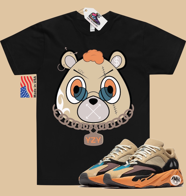 May include: Black t-shirt featuring a cartoon bear wearing a chain and a tag that reads "YZY". The bear has a stitched-up face with a dollar sign on its ear. A pair of tan, orange, and blue trainers with a black sole and a tag that says "1MN" on the side.