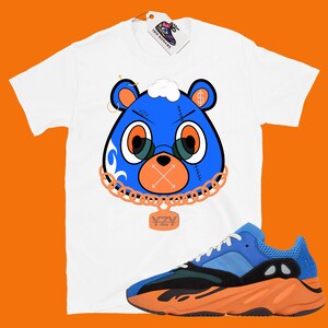 May include: White t-shirt with a blue cartoon bear wearing a gold chain and a 'YZY' pendant. The bear has a dollar sign on its head and a white cloud on top. The t-shirt is paired with blue and orange sneakers with a black midsole.