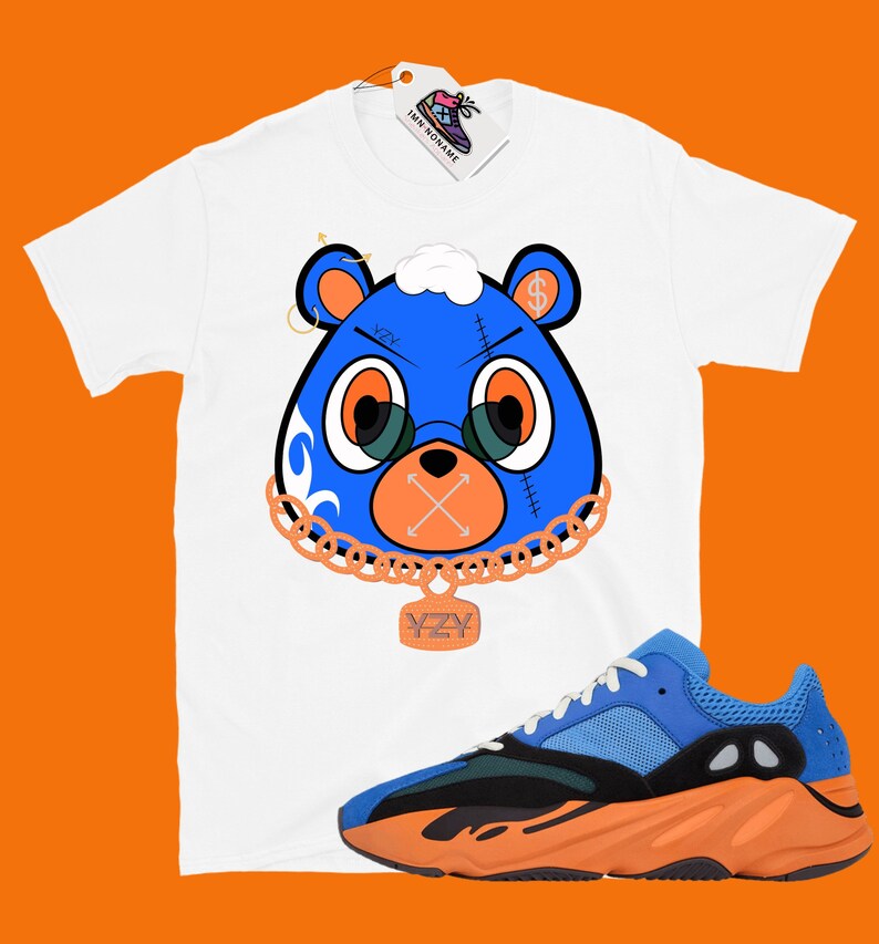 May include: White t-shirt with a blue cartoon bear wearing a gold chain and a 'YZY' pendant. The bear has a dollar sign on its head and a white cloud on top. The t-shirt is paired with blue and orange sneakers with a black midsole.