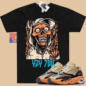 May include: Black t-shirt with a cartoon illustration of a skull wearing a hoodie and making a hand gesture. The text "424 700" is printed below the illustration. A pair of tan, orange, and blue sneakers are shown below the t-shirt.
