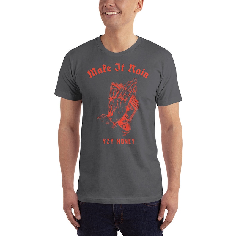Puede incluir: Una camiseta gris oscuro con un gr&aacute;fico rojo de una mano sosteniendo dinero con el texto "Make It Rain" y "YZY MONEY" impreso en la parte delantera.