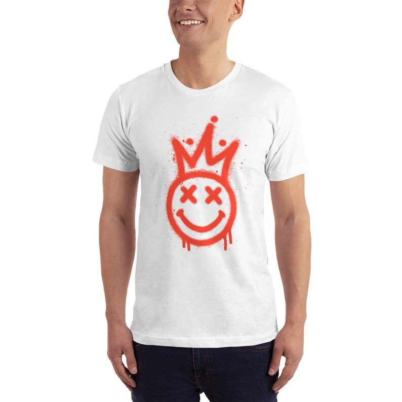 May include: White t-shirt featuring a red spray paint design of a smiley face with a crown on top. The smiley face has X's for eyes.