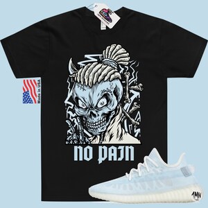 May include: Black t-shirt with a graphic of a skull with dreadlocks and the text "no pain". A pair of white and light blue sneakers are shown below the t-shirt.
