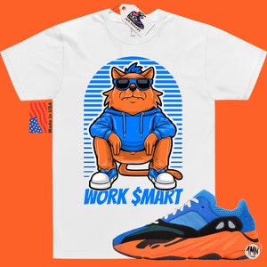 Op de afbeelding: Wit T-shirt met een cartoonkat die een zonnebril en een blauwe hoodie draagt. De kat zit met de tekst "WORK $MART" eronder. Een paar oranje en blauwe sneakers bevinden zich in de rechterbenedenhoek van de afbeelding.