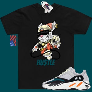May include: Black t-shirt featuring a cartoon rabbit wearing sunglasses and a hat, holding a microphone and spray paint can. The text "HUSTLE" is printed below the rabbit. A pair of white, grey, orange, and teal trainers are shown below the t-shirt.