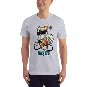 May include: A grey t-shirt featuring a cartoon rabbit wearing a hat, sunglasses and a gold chain. The rabbit is holding a microphone and a spray can. The text "HU$TLE" is printed below the rabbit.