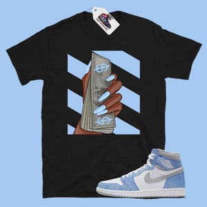 May include: Black t-shirt featuring a graphic of a hand holding a stack of money with blue dollar signs and light blue nails. The background is white with black diagonal stripes. A pair of light blue and white trainers are also pictured.