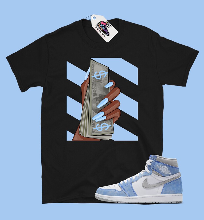 May include: Black t-shirt featuring a graphic of a hand holding a stack of money with blue dollar signs and light blue nails. The background is white with black diagonal stripes. A pair of light blue and white trainers are also pictured.