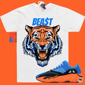 May include: White t-shirt with a tiger graphic and the word "BEA$T" in blue. A pair of blue, black, and orange sneakers is in the foreground. The shirt has a "Made in USA" tag.
