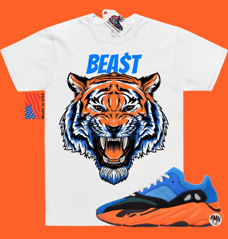 May include: White t-shirt with a tiger graphic and the word "BEA$T" in blue. A pair of blue, black, and orange sneakers is in the foreground. The shirt has a "Made in USA" tag.