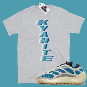 Może przedstawiać: Szary T-shirt z nadrukiem słowa "KYANITE" pionowo w niebieskich i białych literach. Pod T-shirtem widoczna jest para białych i niebieskich but&oacute;w sportowych z czarną podeszwą.