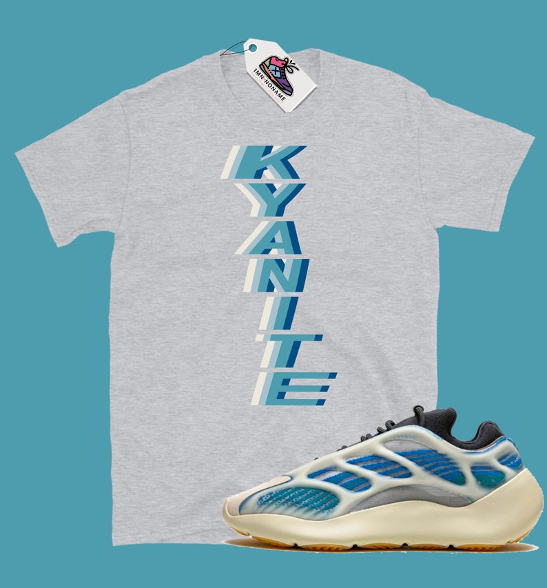 Może przedstawiać: Szary T-shirt z nadrukiem słowa "KYANITE" pionowo w niebieskich i białych literach. Pod T-shirtem widoczna jest para białych i niebieskich but&oacute;w sportowych z czarną podeszwą.