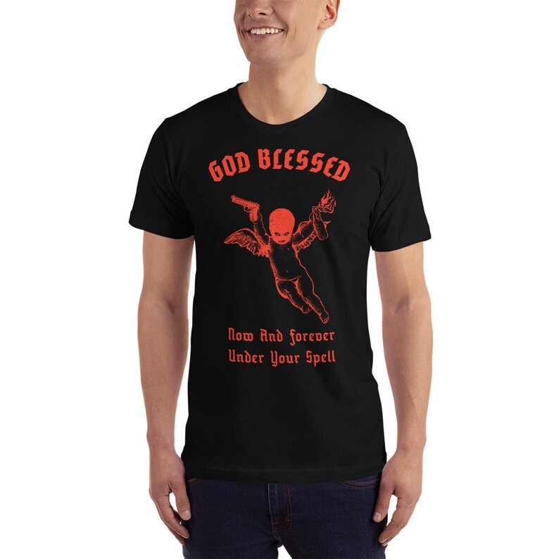 Puede incluir: Camiseta negra con un gr&aacute;fico rojo de un querub&iacute;n sosteniendo una pistola y un c&oacute;ctel Molotov. El texto "GOD BLESSED" est&aacute; por encima del querub&iacute;n y "Now And Forever Under Your Spell" est&aacute; por debajo.