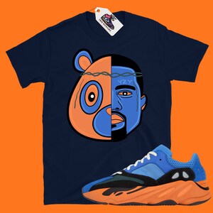 Pode incluir: Uma camiseta azul marinho com um gr&aacute;fico de um urso e o rosto de Kanye West. O gr&aacute;fico &eacute; dividido ao meio, com o urso &agrave; esquerda e Kanye &agrave; direita. O urso &eacute; laranja e azul, e Kanye &eacute; azul. A camiseta &eacute; combinada com t&ecirc;nis laranja e azul.