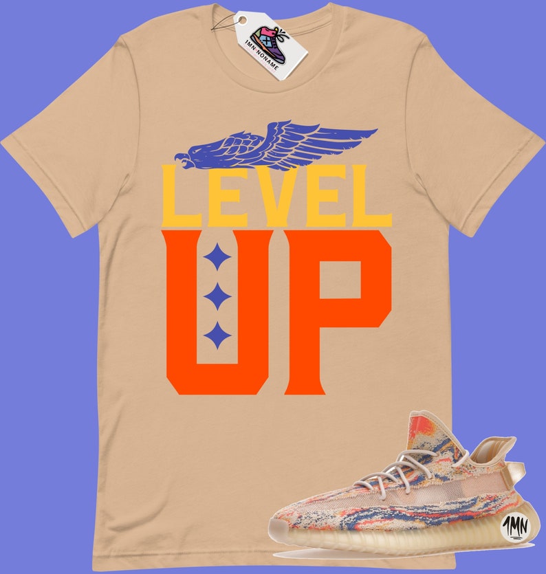 K&ouml;nnte beinhalten: Ein beiges T-Shirt mit den Worten "LEVEL UP" in orangefarbener und blauer Schrift. Ein stilisierter Adler mit blauen Fl&uuml;geln befindet sich &uuml;ber dem Text. Ein Paar beige und mehrfarbige Turnschuhe befinden sich in der rechten unteren Ecke des Bildes.