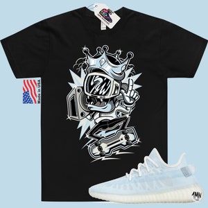 Puede incluir: Camiseta negra con una ilustraci&oacute;n de dibujos animados de un personaje que lleva una corona y sostiene una radio. El personaje est&aacute; de pie sobre una patineta. La camiseta se combina con un par de zapatillas de deporte azul claro con suela blanca.