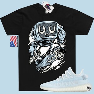 May include: Black t-shirt with a cartoon illustration of a robot with a television for a head skateboarding. The robot is wearing a blue jumpsuit and has a white cloud behind it. The t-shirt has the text "Made in USA" on the left side. A pair of white and light blue sneakers with the text "1MN" on the side are shown below the t-shirt.
