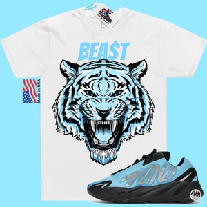 以下が含まれることがあります： 青い虎のグラフィックと青い文字で「BEAST」と書かれた白いTシャツ。側面に「700」と記された黒と水色のスニーカーのペア。