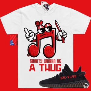 Puede incluir: Camiseta blanca con una nota musical roja de dibujos animados que sostiene un cuchillo y el texto "Shorty Wanna Be A Thug". Zapatillas negras y rojas con el texto "YZY-350" en el lateral.